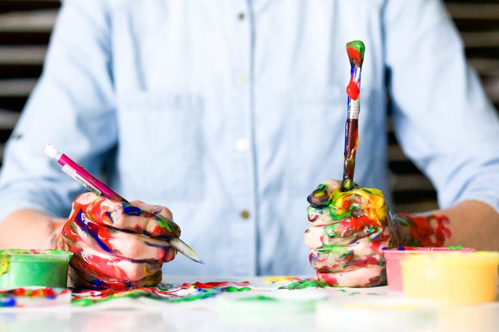 Man painting at table with hands and painbrush covered in different colors of paint