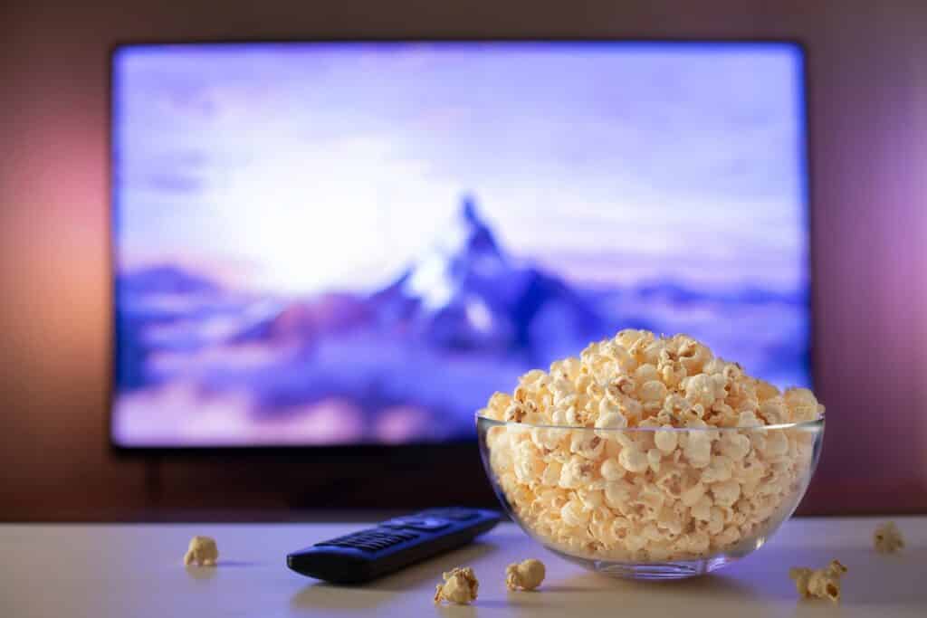 A glass bowl of popcorn and remote control in the background the TV works. Evening cozy watching a movie or TV series at home.