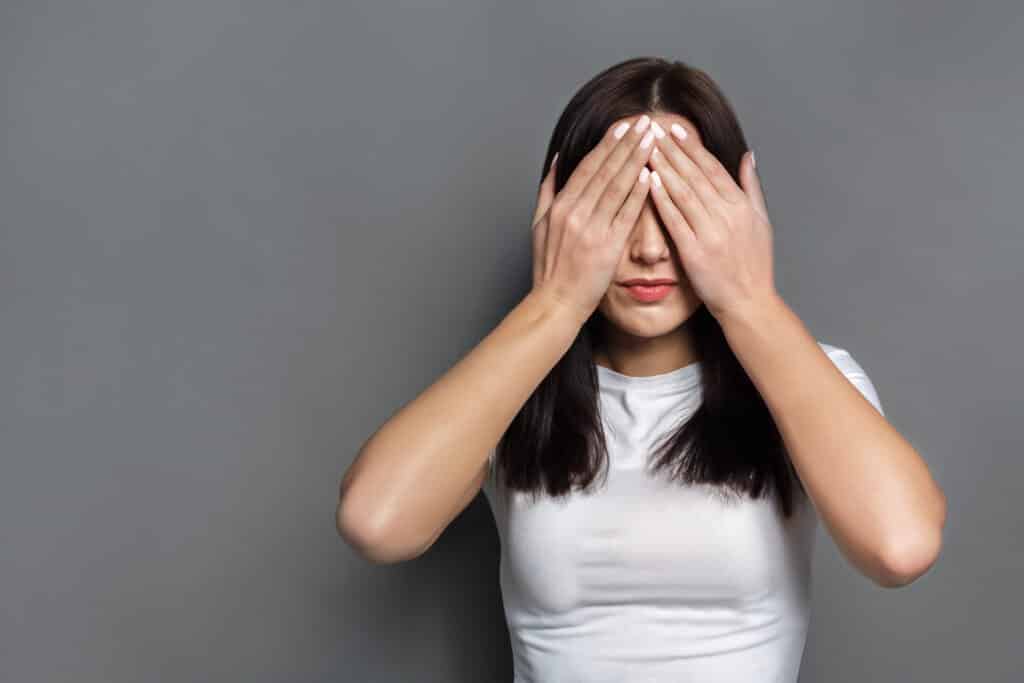 See no evil concept. Portrait of young scared woman covering eyes with hands while standing against gray studio background. Confused girl close eyes with palms ignoring something